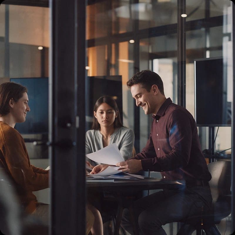 Three colleagues in a glass-walled meeting room reviewing papers together.